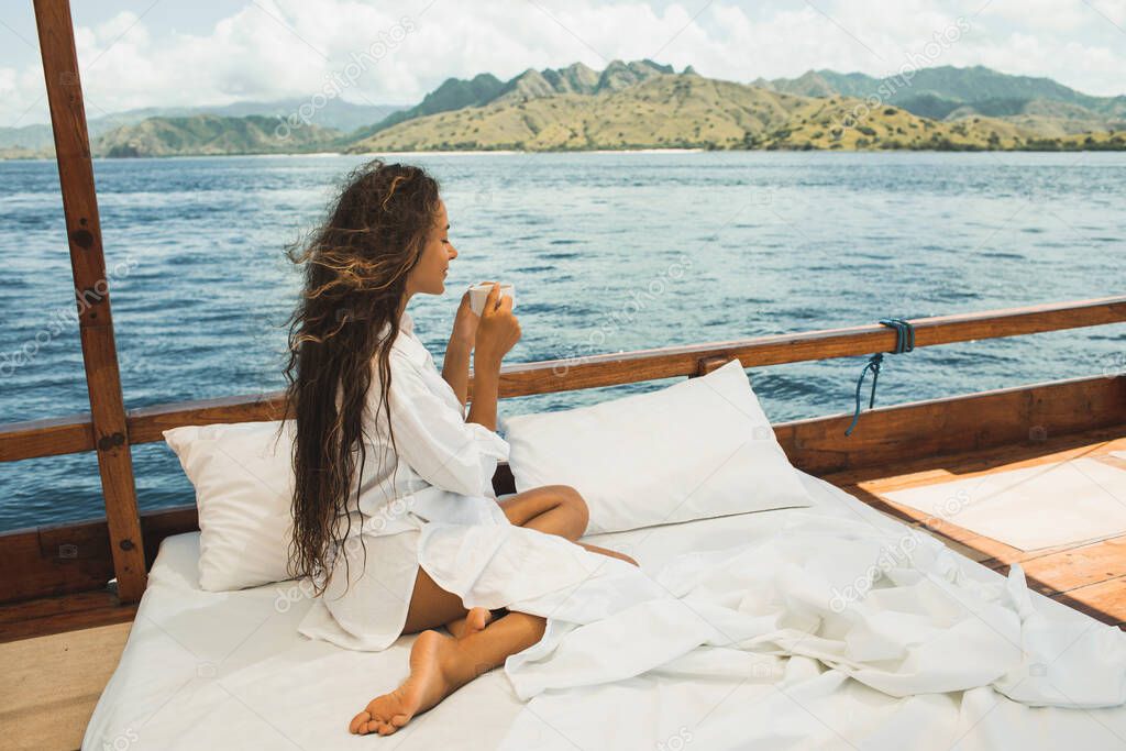 Woman enjoying morning coffee on boat in private cruise tour. Bed on board. Travel and freedom lifestyle. Mountain view of Komodo national park on horizon.