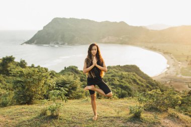 Gün batımında yoga yapan genç bir kadın güzel bir okyanus ve dağ manzarasıyla poz veriyor. Doğaya karşı duyarlılık. Aktif yaşam tarzı. Ruhani ve duygusal kavram.