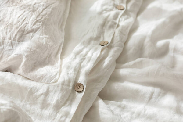 Trendy organic natural linen bedclothes with wooden buttons closeup. Bedding, morning light, bedroom style and design. Rough textile background with wrinkle.