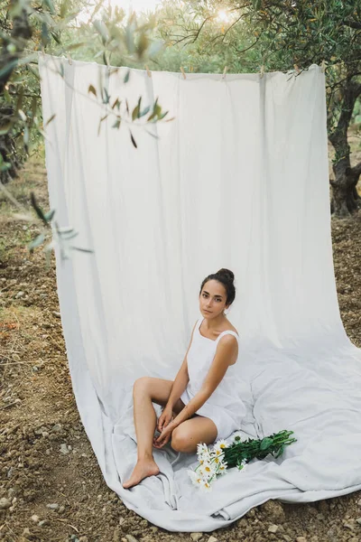 テキスタイルの布をぶら下げの背景に白い夏のリネンのドレスで若いブルネットの女性の肖像画 リラクゼーションとレジャー オリーブの庭でピクニック カモミールの花の花束 — ストック写真
