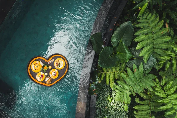 Floating breakfast in jungle swimming pool, tropical resort. Black rattan tray in heart shape, Valentines day or honeymoon surprise, view from above. Exotic summer diet. Tropical beach lifestyle.