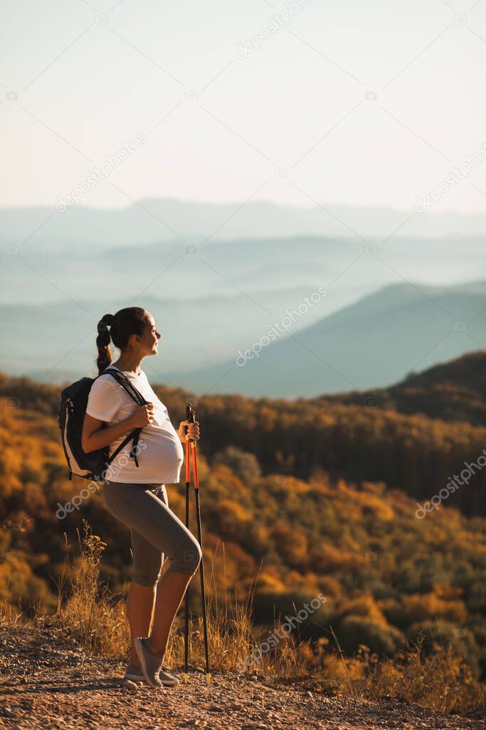 Pregnant woman nordic walking with trekking sticks on hill with beautiful autumn mountain view. Pregnancy activity and healthy lifestyle in maternity time. Travel concept.
