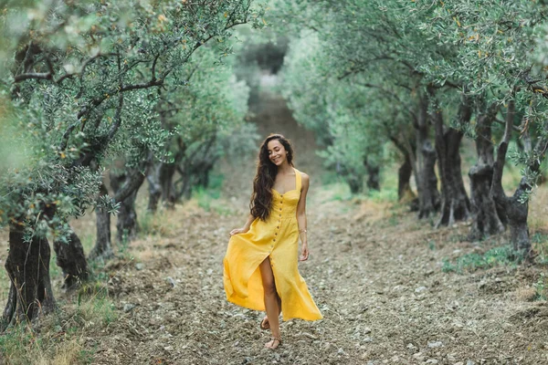 若い幸せな笑顔の女性のオリーブの木の庭を歩く 黄色のリネン夏のドレス 流行の色と繊維のスタイル — ストック写真