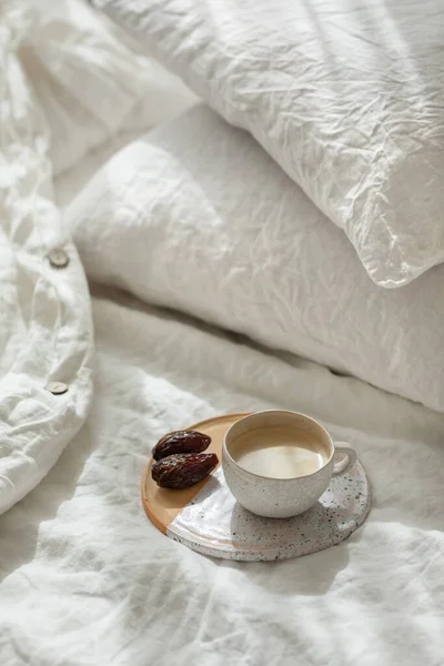 Cup of fresh coffee in bed on ceramic tray, morning mood. Linen cotton textile bedclothes. Organic and natural linen. Cozy bedroom interior. Beautiful light.