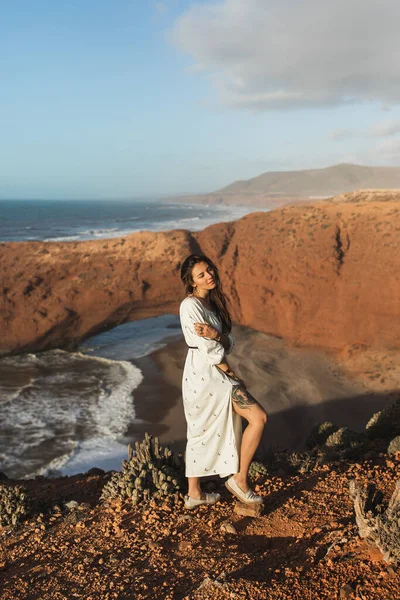 モロッコのLegziraビーチを背景に白いドレスで若いスタイリッシュな美しい女性の肖像画 裸の脚の入れ墨 — ストック写真