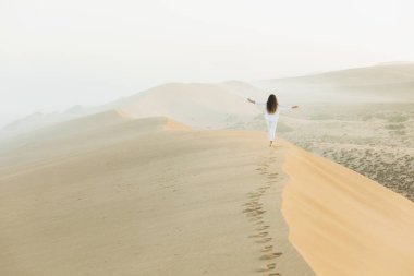 Fas Sahra Çölü 'nde kumulun tepesinde yürüyen bir kadın. Güzel, sıcak güneş ışığı ve sabah sisi. Arkadan bak. Özgürlük kavramı.
