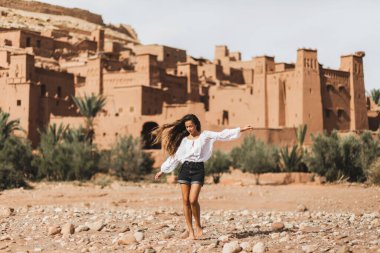 Genç kıvırcık kadın doğuda Kasbah Ait-Ben-Haddou 'nun arka planında yürüyor. Fas 'a git, Ouarzazate. Yaz tatilleri, seyahat yaşam tarzı konsepti..