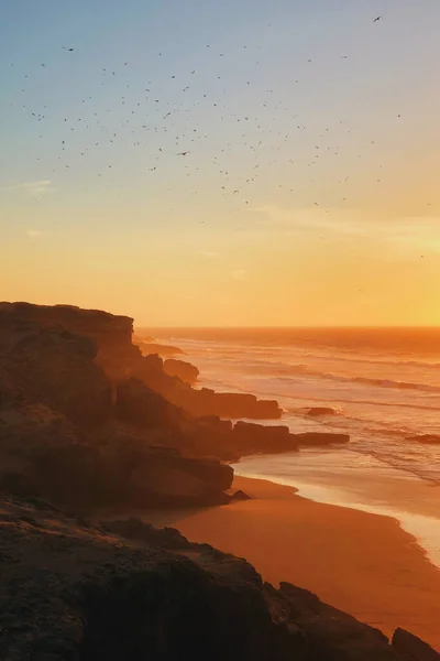 Fantastic orange sunset with a lot of flying birds. Layered coastline with water mist near Agadir, Morocco.