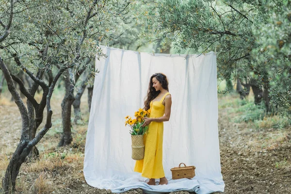 庭で白いハングテキスタイル布を背景に若いブルネットの女性の肖像画 黄色の花 夏のリネンのドレスとわらバスケットを保持 — ストック写真