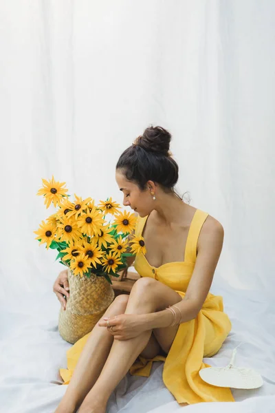 若いブルネットの女性の肖像画黄色の花 夏のリネンのドレスの藁バスケットと白い吊り織物の布を背景に — ストック写真
