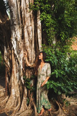 Tropikal bahçede yeşil elbiseli güzel Asyalı kadın portresi. Güzel gün batımı ışığı, büyük ağaç, inanılmaz doğa..
