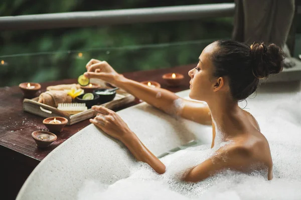 Banyo yapan bir kadın. Köpüklü küvet. Romantik spa tedavisi, vücut bakımı. Doğal kozmetik ürünleri ahşap tepside..