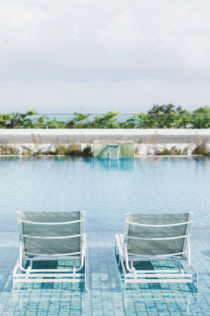 White metal recliners on pool side. Contemporary beach outdoor furniture. Swimming pool with clear blue water. Luxury vacations in Asia.