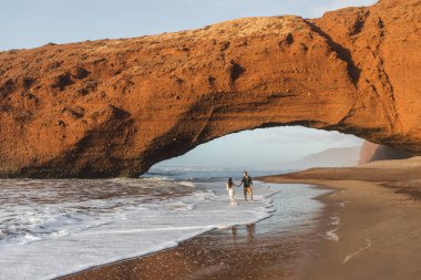 Couple in love walking under huge arch on Legzira ocean beach at sunset. Famous and popular landmark in Morocco. clipart