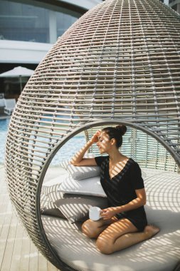 Genç Asyalı kadın dışarıda kozası şeklinde kahve içiyor ve dinleniyor. Rattan 'dan yapılmış. Dış mobilyaların çağdaş tasarımı.