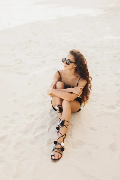 年轻的黑发女子 戴着太阳镜 身穿黑色泳衣 在海滩上放松 日落时坐在白沙滩上 — 图库照片