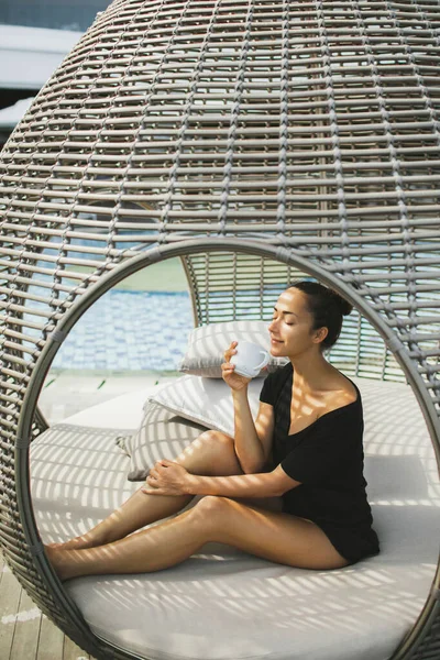 年轻的亚洲女人在户外的床上放松地喝咖啡 长成茧状 用藤条做的室外家具的当代设计 — 图库照片