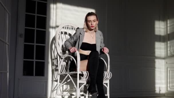 Mujer de moda en la chaqueta se sienta en un sillón blanco en la habitación — Vídeos de Stock