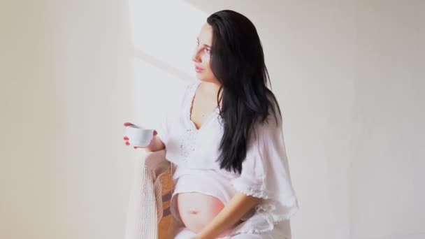 En gravid kvinna med en kopp te ser ut genom fönstret — Stockvideo