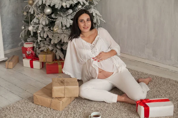 A pregnant woman sitting by the Christmas tree opens Christmas presents — Stock Photo, Image