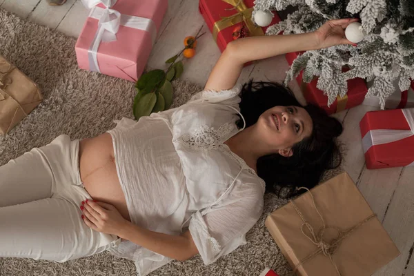 A pregnant woman near a Christmas tree opens Christmas presents — Stock Photo, Image