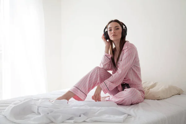 Mooie vrouw in roze pyjama's, luisteren naar muziek met een koptelefoon op het bed — Stockfoto