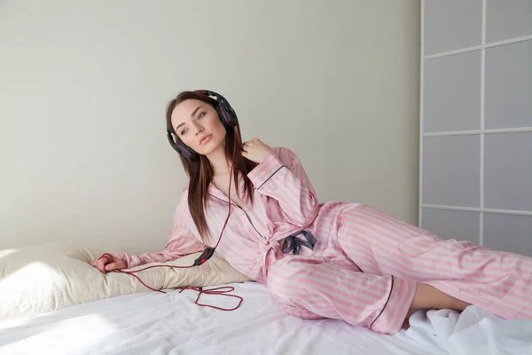 ピンクのパジャマ、ベッドの上にヘッドフォンで音楽を聴くには美人 — ストック写真