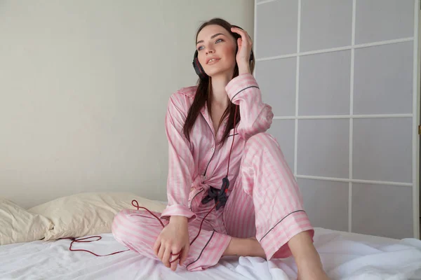 Mooie vrouw in roze pyjama's, luisteren naar muziek met een koptelefoon op het bed — Stockfoto