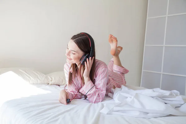 Mooie vrouw in roze pyjama's, luisteren naar muziek met een koptelefoon op het bed — Stockfoto