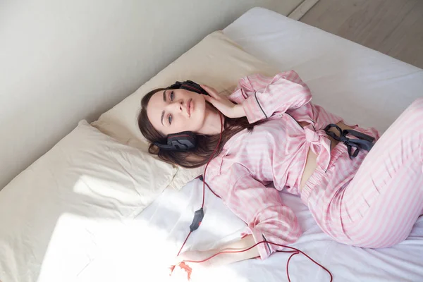 Mooie vrouw in roze pyjama's, luisteren naar muziek met een koptelefoon op het bed — Stockfoto