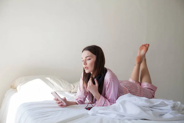 Mooie vrouw in roze pyjama's, luisteren naar muziek met een koptelefoon op het bed — Stockfoto