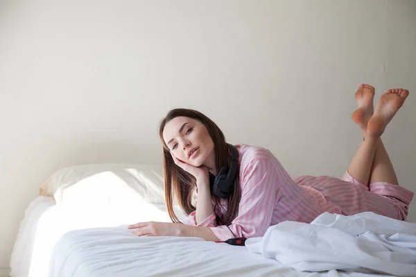 Mooie vrouw in roze pyjama's, luisteren naar muziek met een koptelefoon op het bed — Stockfoto