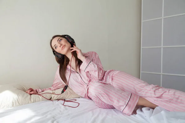 Женщина в розовых пижамах слушает музыку в наушниках, танцующих на кровати — стоковое фото