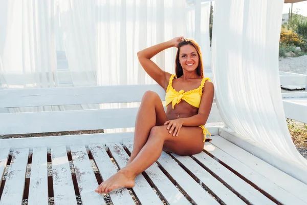 Opalona kobieta w żółtym stroju kąpielowym z długimi włosami siedzi w cieniu na plaży — Zdjęcie stockowe