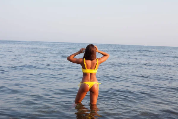 Beautiful tanned woman in yellow swimsuit stands in the sea bathing on the beach — Stock Photo, Image