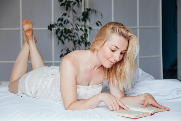 Hermosa mujer rubia lee un libro en el dormitorio en la cama —  Fotos de Stock