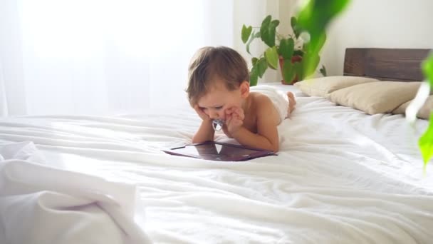 Niño pequeño mira en la tableta acostado en la cama — Vídeos de Stock