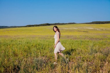 Elbiseli güzel bir kadın sarı çiçeklerle sahada yürüyor.