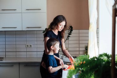 Küçük oğlu annesine sebze pişirmede yardım ediyor. Mutfakta sebze doğruyor.