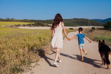Anne ve oğlu sarı tarlada köpekle yürüyor.