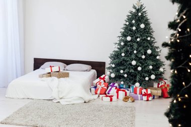Beyaz yatak odasının içinde mavi Noel ağacı olan bir yatak ve yeni yıl için kırmızı hediyeli kış kartpostalı.