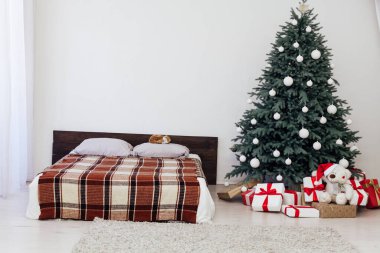 Beyaz yatak odasının içinde mavi Noel ağacı olan bir yatak ve yeni yıl için kırmızı hediyeli kış kartpostalı.