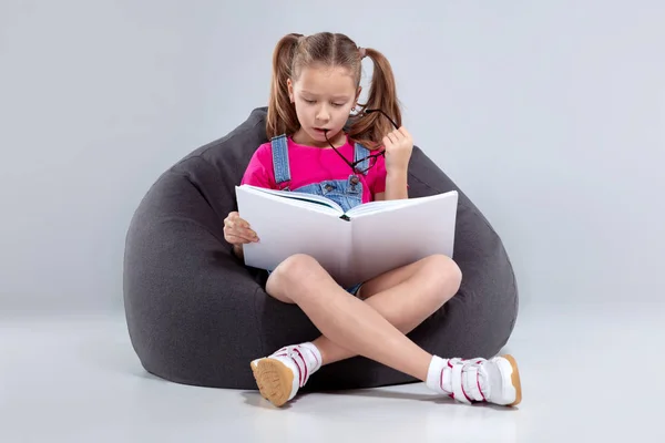 Mladá dívka v brýlích čtení knihy na šedou sedací žok — Stock fotografie