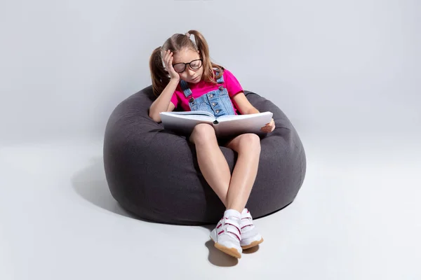 Chica joven en gafas durmiendo con un libro en una bolsa de frijoles grises —  Fotos de Stock