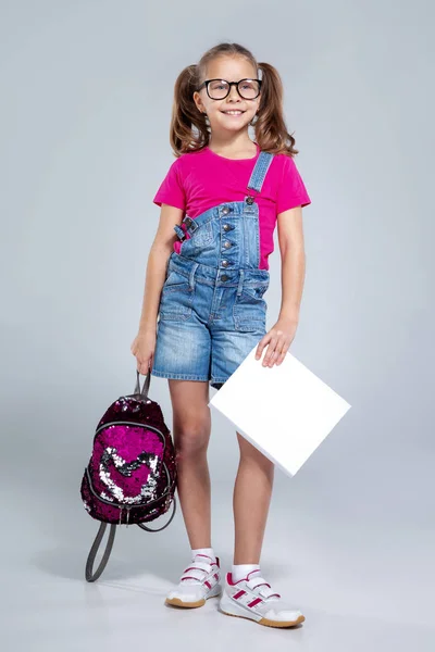 Colegiala en gafas con mochila y libro de texto sobre el fondo gris — Foto de Stock