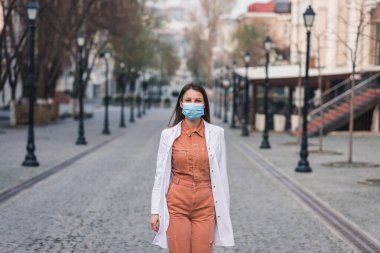 Turuncu elbiseli esmer bir kadın beyaz bir ceket giyiyor ve yüzünde koruyucu bir maske var.