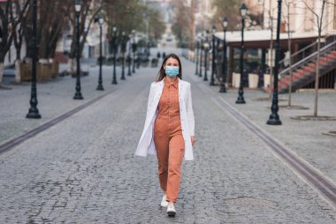 Turuncu elbiseli esmer bir kadın beyaz bir ceket giyiyor ve yüzünde koruyucu bir maske var.