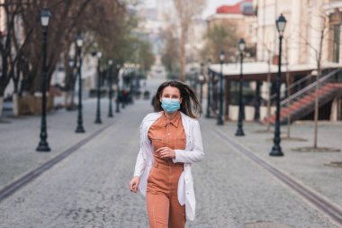 Turuncu elbiseli esmer bir kadın beyaz bir ceket giyiyor ve yüzünde koruyucu bir maske var.