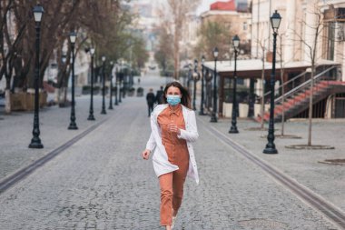 Turuncu elbiseli esmer bir kadın beyaz bir ceket giyiyor ve yüzünde koruyucu bir maske var.