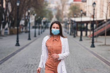 Turuncu elbiseli esmer bir kadın beyaz bir ceket giyiyor ve yüzünde koruyucu bir maske var.